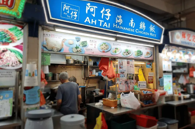 An eye-level shot framing the bustling interior of the Ah Tai Hainanese Chicken Rice stall, showing a vendor working among stainless steel pots beneath a bright blue and white storefront sign.