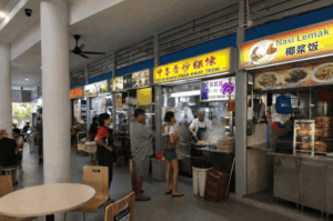 A bustling hawker centre where customers queue at food stalls. Inside a Char Kway Teow stall, a chef cooks over a hot wok, sending a thick plume of steam and smoke into the air.