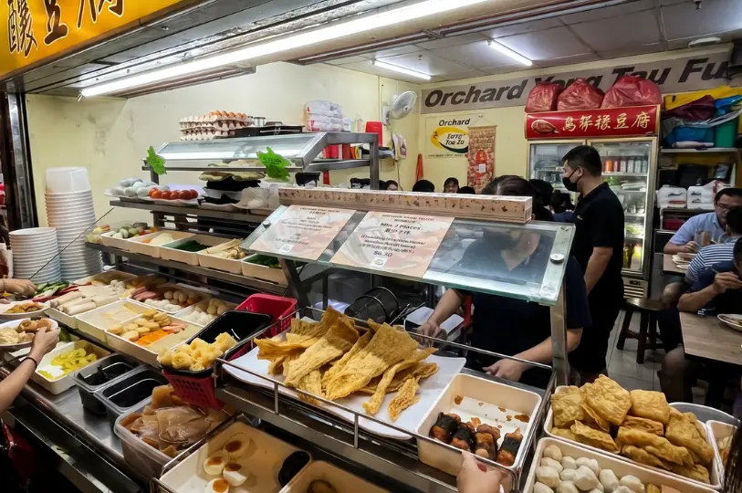 Customers selecting their meal from a wide, colorful assortment of raw vegetables, fishballs, stuffed tofu, and crispy fried bean curd skins at the self-serve counter.
