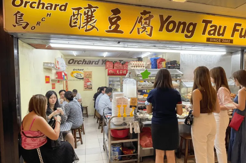 A bustling scene at Orchard Yong Tau Fu, featuring its bright yellow signboard and a line of customers selecting ingredients, with other patrons eating at small tables in the compact stall.