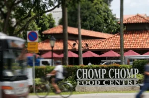 A blurred bus and cyclist speed past the large entrance sign for Chomp Chomp Food Centre, featuring a distinctive red-tiled roof and an outdoor dining area shaded by red umbrellas.
