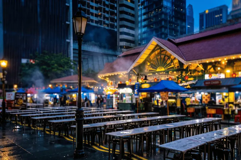 Rows of empty outdoor tables and stools reflecting on a wet street at dusk, set up outside the warmly lit Victorian-style Lau Pa Sat market.