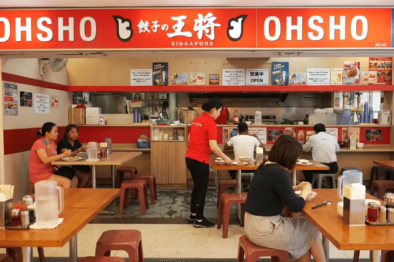 Customers dining at wooden tables with red plastic stools at Gyoza no Ohsho, a casual Japanese eatery with a prominent red and white signboard and staff members in matching red uniforms.