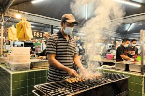 A hawker stall worker wearing a face mask grilling skewers over hot coals at a busy Singapore food market.