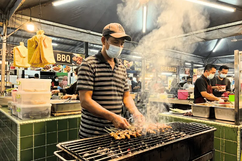 A hawker stall worker wearing a face mask grilling skewers over hot coals at a busy Singapore food market.