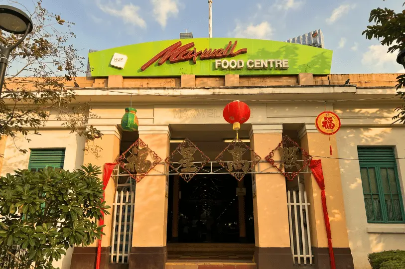 The exterior facade of Maxwell Food Centre bathed in warm sunlight, showcasing the distinctive green arched rooftop sign and the main entrance decorated with festive red lanterns and Chinese banners.