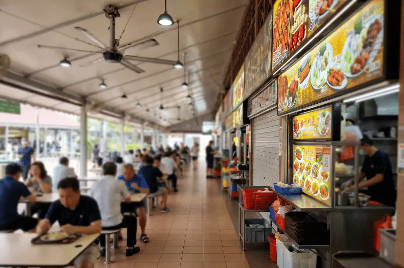 An aisle-perspective shot with a shallow depth of field, leading the eye past brightly lit menu boards on the right toward softly focused diners sitting at tables under large ceiling fans on the left.