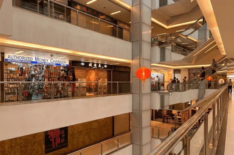 The multi-level interior atrium of Cuppage Plaza, featuring escalators connecting the floors and red decorative lanterns hanging in the open space. The walkways are lined with a diverse mix of tenants, including a brightly lit money changer packed with snacks positioned right next to a traditional Japanese restaurant, capturing the eclectic, bustling atmosphere of Singapore's Little Tokyo.