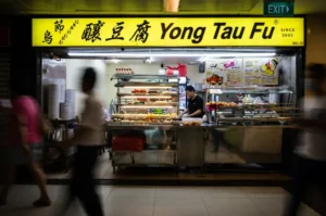 The bright yellow storefront of Orchard Yong Tau Fu in Cuppage Plaza, with a worker preparing food behind a glass display of ingredients as blurred pedestrians walk by.