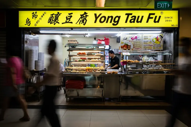 The bright yellow storefront of Orchard Yong Tau Fu in Cuppage Plaza, with a worker preparing food behind a glass display of ingredients as blurred pedestrians walk by.