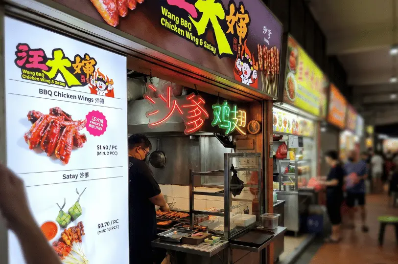 A vendor in a black shirt cooks skewers over a smoky grill at the brightly lit Wang BBQ Chicken Wing & Satay stall, which features glowing neon signage and a large, illuminated menu board.