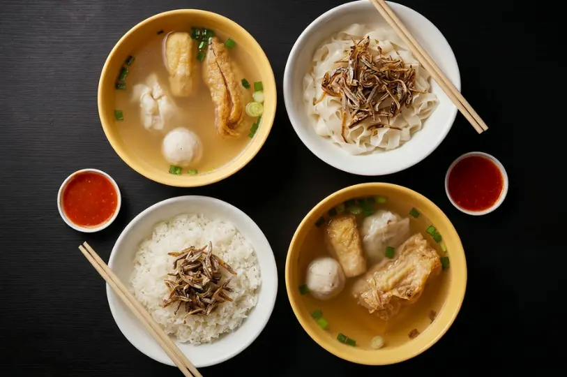 An overhead view of a hearty Yong Tau Fu meal set on a dark table, featuring bowls of soup with stuffed tofu and fishballs, alongside portions of flat noodles and white rice topped with crispy anchovies.