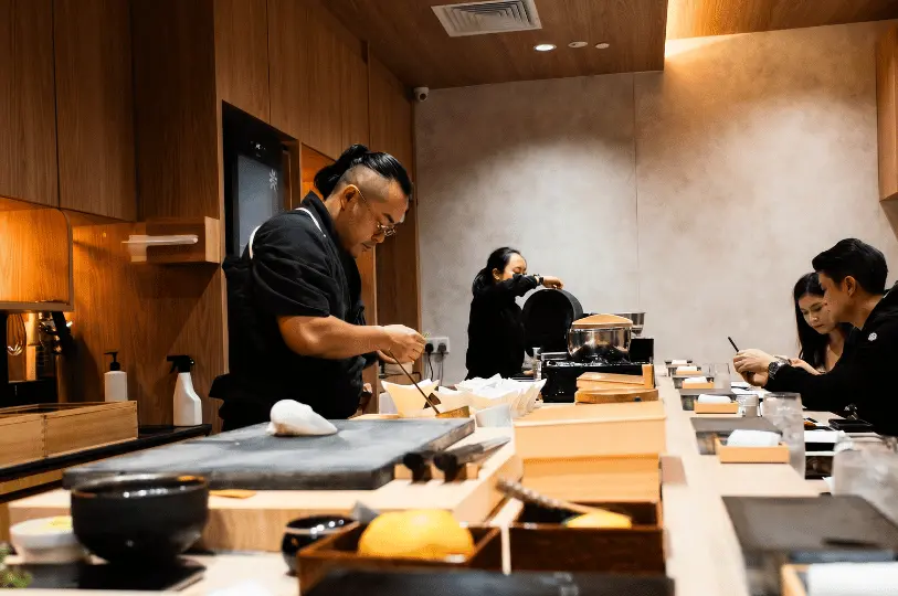 A chef with a tied-back hairstyle focuses on preparing a dish behind a brightly lit, minimalist wooden sushi counter, while a couple sits quietly at the counter waiting for their meal.