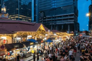 Wide evening shot of the bustling Lau Pa Sat Satay Street, showing crowds dining outdoors among glowing food stalls, with the historic clock tower and modern skyscrapers in the background.