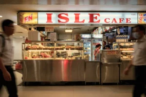 A front view of the Isle Cafe food stall featuring a brightly lit sign with red text above stainless steel display counters filled with various foods, while blurred figures of pedestrians walk past in the foreground.