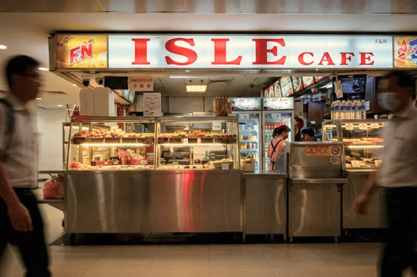 A front view of the Isle Cafe food stall featuring a brightly lit sign with red text above stainless steel display counters filled with various foods, while blurred figures of pedestrians walk past in the foreground.