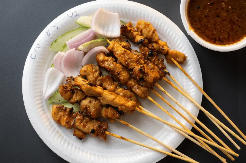 A close-up, overhead view of grilled chicken satay skewers on a white disposable plate, garnished with thick slices of raw cucumber and red onion, served next to a bowl of chunky peanut dipping sauce.