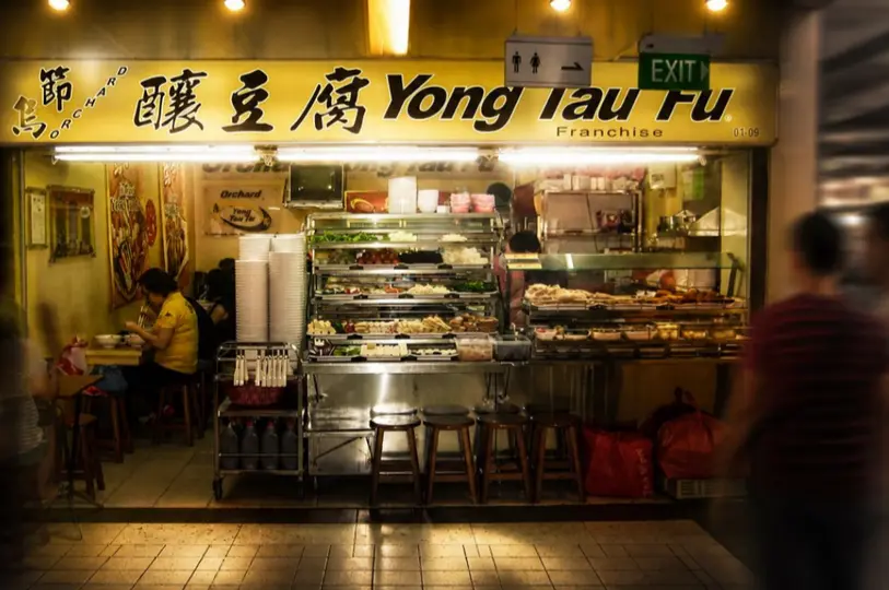 A warm, slightly moody view of the Orchard Yong Tau Fu stall showing a diner eating quietly at a small table next to the bustling ingredient counter under the glowing yellow signboard.