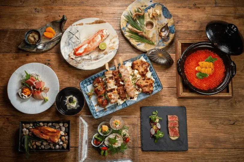 A top-down view of an elaborate Japanese feast arranged on a rustic wooden table, showcasing a variety of dishes including yakitori skewers, fresh sashimi, a crab leg, and an earthenware pot filled with bright orange ikura (salmon roe).