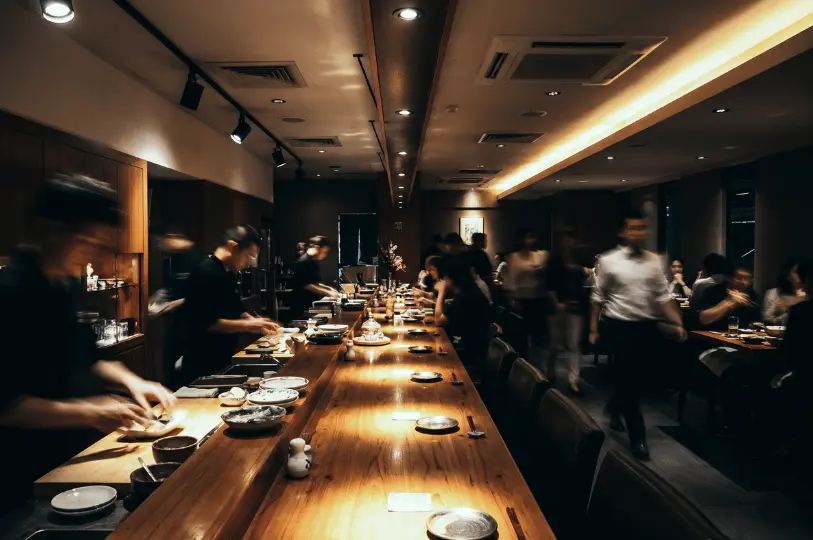 A dimly lit Japanese restaurant interior showing a long, warmly illuminated wooden counter. Chefs prepare food while patrons dine, captured with motion blur to convey the bustling atmosphere without focusing on individual faces.