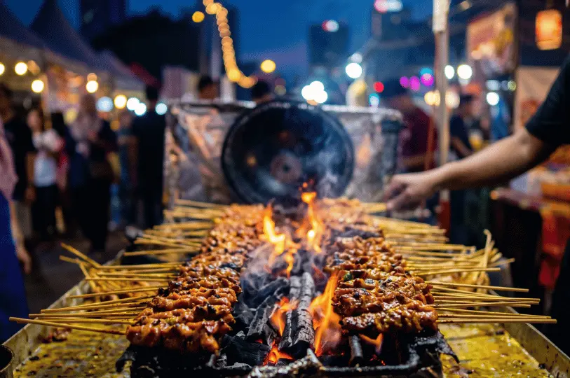 Close-up of dozens of satay skewers grilling over a smoky, open-flame charcoal pit, with the bustling night market blurred in the background.