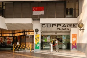 The exterior entrance of Cuppage Plaza in Singapore, featuring its name sign above glass doors, a hanging Singapore flag, and the storefront of Shinjuku Japanese restaurant to the left.