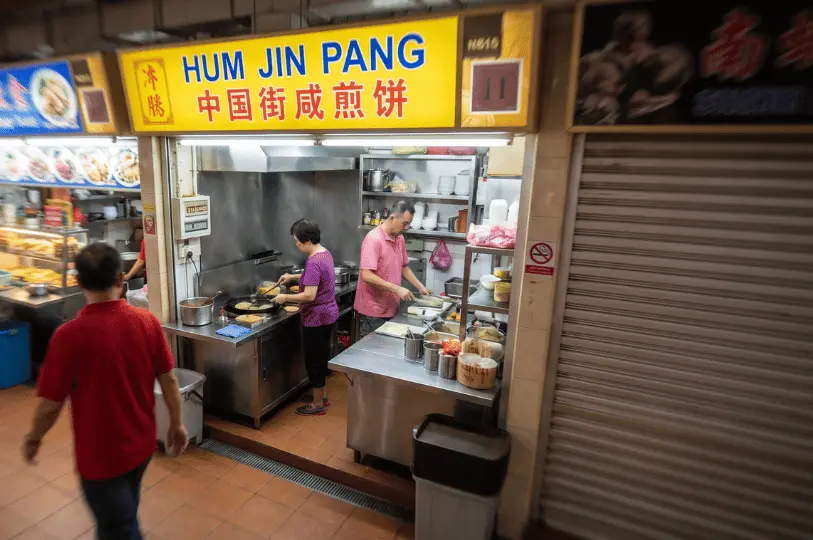 A candid, slightly elevated shot of two hawkers actively cooking inside the brightly lit Hum Jin Pang stall, contrasted with a motion-blurred pedestrian in a red shirt walking through the foreground.