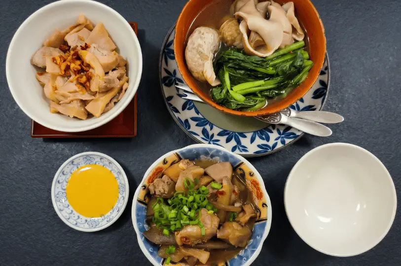 An overhead shot of a home-style Asian meal on a dark textured table, featuring bowls of braised meats, root vegetables, a clear broth with greens and dumplings, and a small dish of yellow dipping sauce.