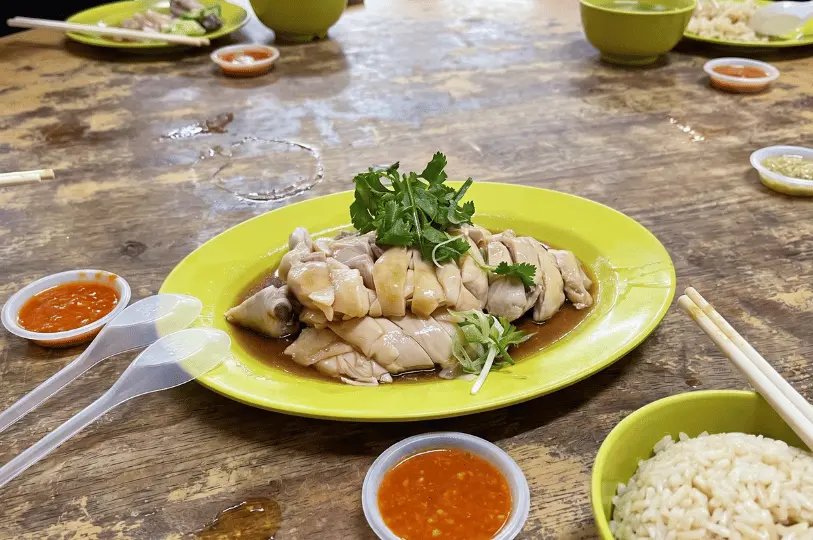 A large, communal plate of sliced poached chicken garnished with fresh cilantro on a rustic wooden table, surrounded by small condiment cups of chili and ginger paste.