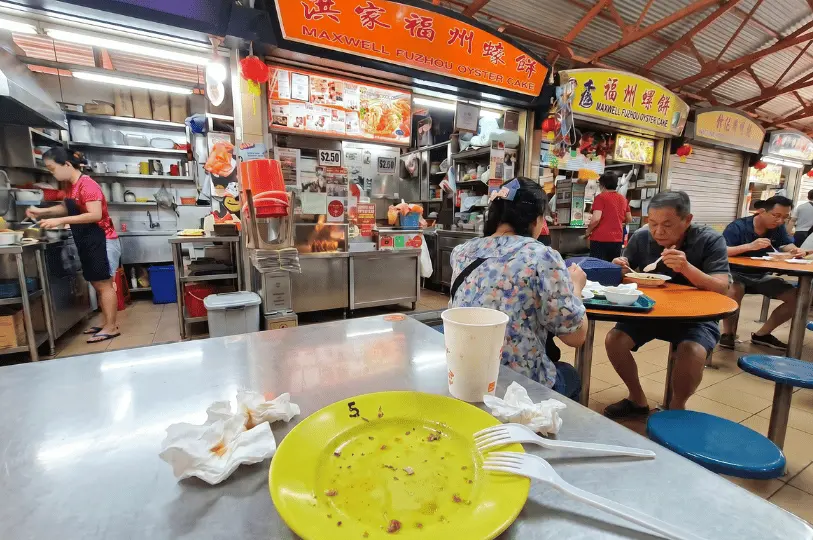 An empty yellow plate with greasy crumbs, crumpled napkins, and plastic forks on a stainless steel table, showing the aftermath of a meal at a busy hawker centre.