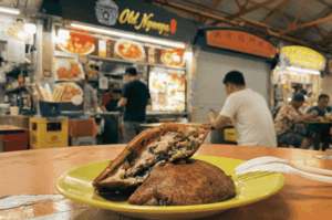 A close-up of a halved, golden-brown, crispy oyster cake on a bright yellow plate. The cross-section reveals a savory, packed filling of minced meat, oysters, and fresh green herbs. It sits on a table in a bustling hawker centre, with patrons and a food stall named "Old Nyonya" visible in the blurred background.