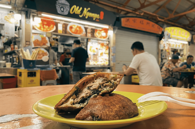 A close-up of a halved, golden-brown, crispy oyster cake on a bright yellow plate. The cross-section reveals a savory, packed filling of minced meat, oysters, and fresh green herbs. It sits on a table in a bustling hawker centre, with patrons and a food stall named "Old Nyonya" visible in the blurred background.