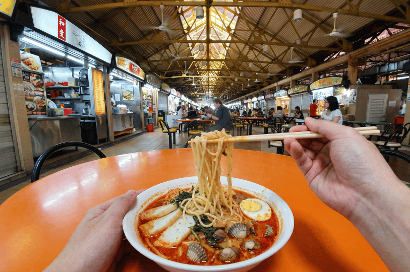 A first-person view of a diner using wooden chopsticks to lift a portion of yellow noodles from a steaming bowl of spicy red noodle soup, likely Laksa. The bowl, resting on a bright orange table, contains fish cake slices, fresh cockles, dark green vegetables, and half a boiled egg. The background shows a bustling hawker centre with food stalls and other patrons dining under a high, yellow-beamed corrugated roof.