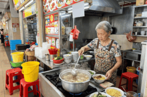 An elderly woman with short gray hair and a floral apron carefully stirs a large, steaming pot of broth with a metal ladle at a hawker stall. She stands behind a stainless steel counter holding bowls of freshly prepared yellow noodles and greens, surrounded by the everyday bustle of colorful plastic buckets, stacked bowls, and red stools.