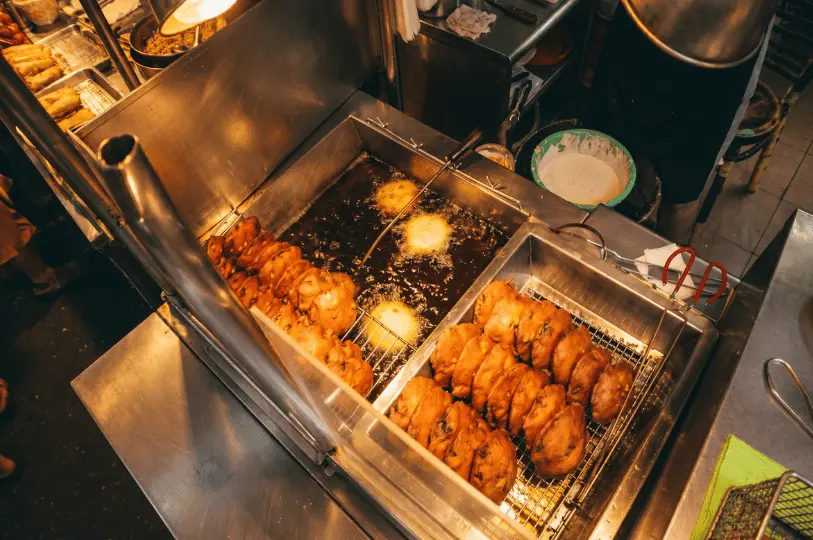 Overhead view of Fuzhou oyster cakes deep-frying in a vat of bubbling hot oil, right next to a tray of finished cakes draining on a metal rack.