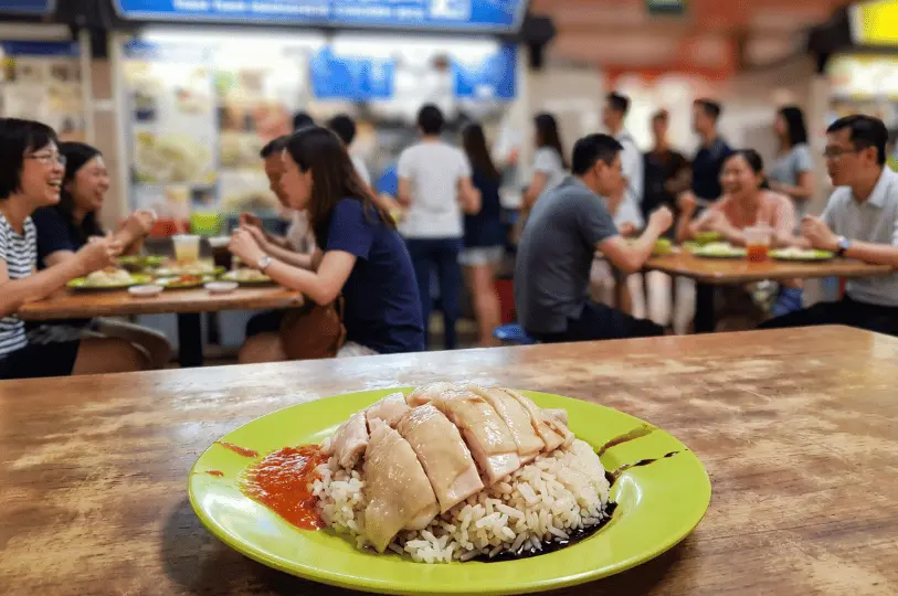 A freshly prepared plate of Hainanese chicken rice with chili sauce on a bright green plate in sharp focus, set against a blurred background of patrons eating at a busy hawker centre.