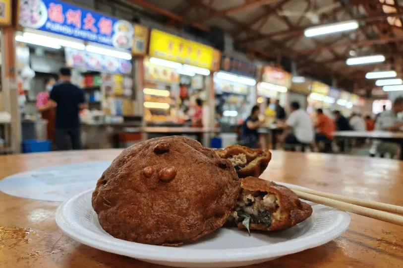 Close-up of crispy Fuzhou oyster cakes on a white disposable plate with wooden chopsticks, one broken open to show the oyster and meat filling, set against a blurred hawker centre background.