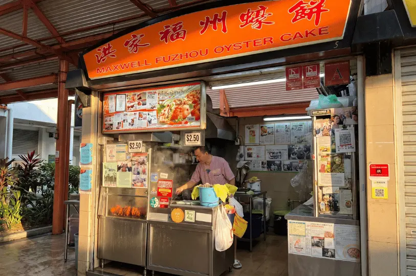 The illuminated storefront of the Maxwell Fuzhou Oyster Cake stall, featuring a bright orange signboard, newspaper clippings, and a vendor actively cooking inside.