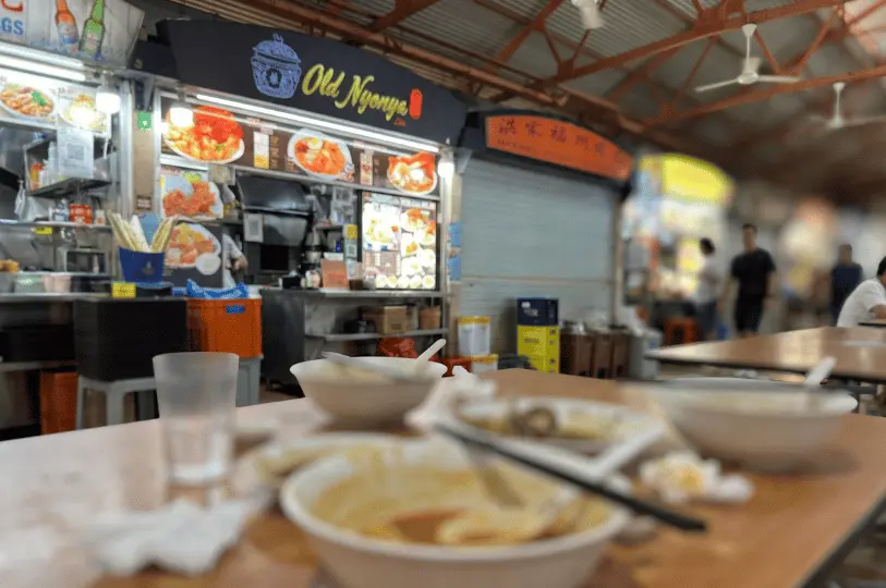 A blurred close-up of empty, messy bowls and crumpled napkins on a table after a finished meal, with the brightly lit Old Nyonya stall visible in the background.
