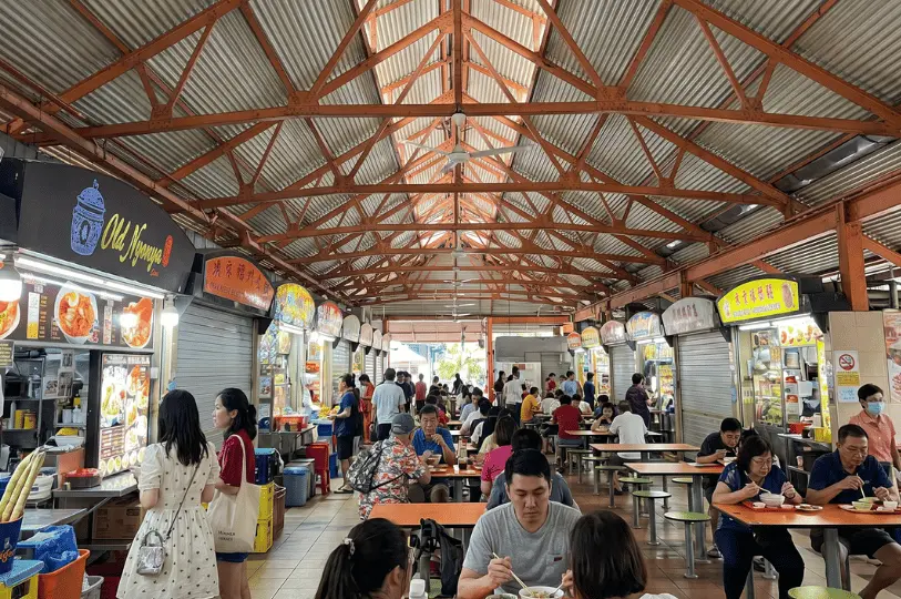 A wide view down the busy central aisle of Maxwell Food Centre. Numerous diners are eating at orange tables under a high, metal roof, with the Old Nyonya stall situated on the left side.
