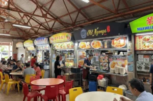 The Old Nyonya food stall at Maxwell Food Centre, featuring a black signboard with gold lettering. In the foreground, diners are eating at round tables with red and yellow chairs.