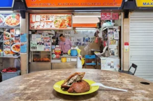 A halved Fuzhou oyster cake on a yellow plate revealing its savory filling, placed on a wooden table with the Maxwell Fuzhou Oyster Cake stall and a vendor in the background.