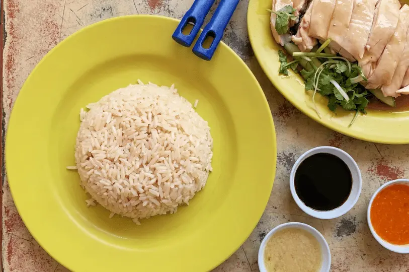 An overhead view of a meal featuring a dome of fragrant rice on a green plate, a separate plate of sliced chicken and cucumber, and trio of small dipping sauces: dark soy, chili, and ginger.