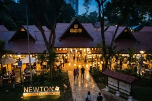 An elevated twilight shot of Newton Food Centre in Singapore, showcasing its iconic orange-tiled roofs, warm ambient lighting, outdoor dining tables, and visitors walking along the central pathway.