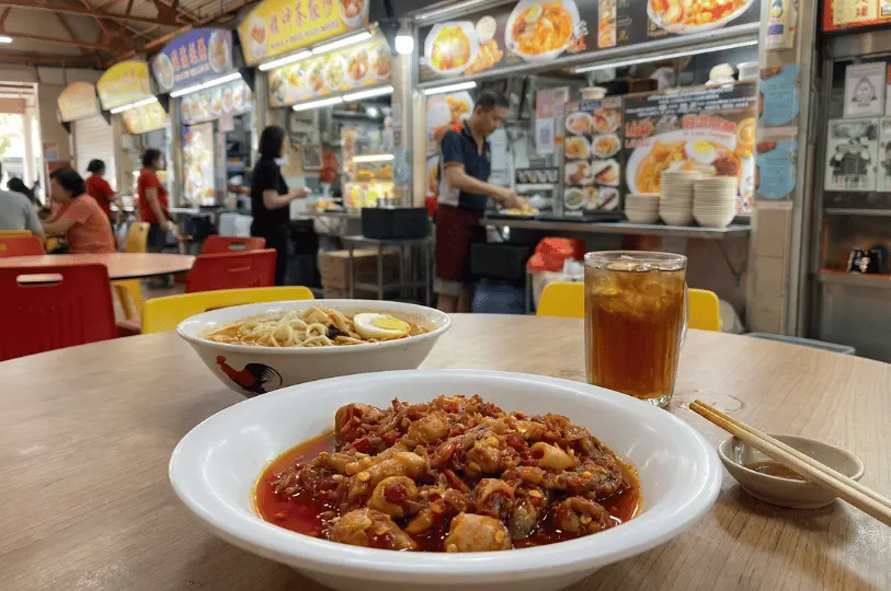 A close-up of a hawker meal on a wooden table, featuring a shallow bowl of vibrant, spicy sambal dish in the foreground and a bowl of laksa behind it, alongside a glass of iced tea. In the softly blurred background, diners eat at bright red and yellow tables while a hawker prepares food at a stall.