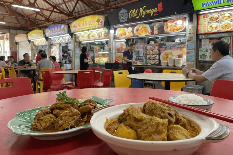A close-up of a plate of chicken rendang and a bowl of chicken curry with potatoes, served with a small bowl of white rice on a red table at Maxwell Food Centre.